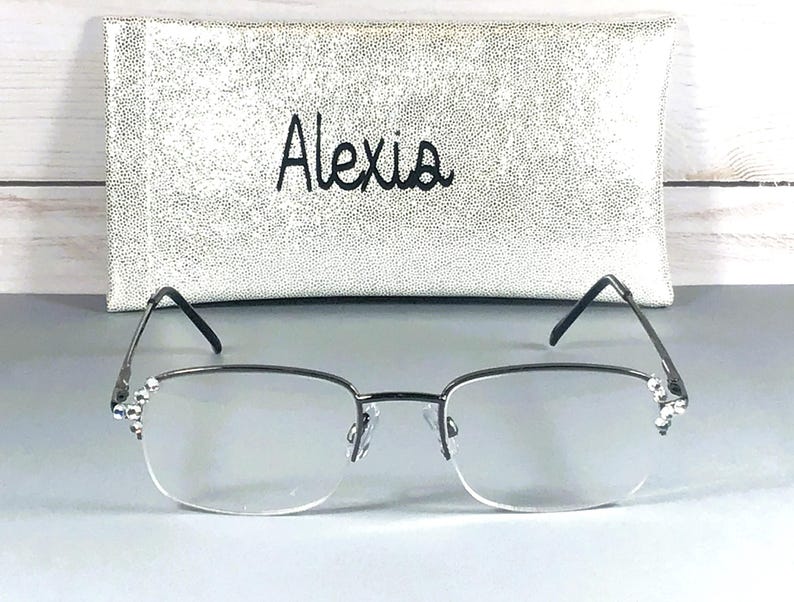 Pode incluir: &Oacute;culos com arma&ccedil;&atilde;o prateada e lentes transparentes, com strass decorativos nas hastes. Atr&aacute;s dos &oacute;culos, uma capa de &oacute;culos prateada e texturizada com o nome "Alexis" bordado em preto. A capa tem um padr&atilde;o de pontos.