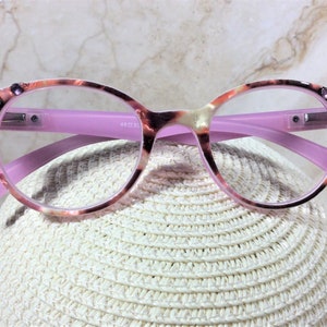 May include: Round-framed eyeglasses with a pink and tortoiseshell pattern. The frames have clear lenses and are accented with rhinestones on the upper corners. The temples are a solid pink color. The glasses are resting on a white, woven surface.
