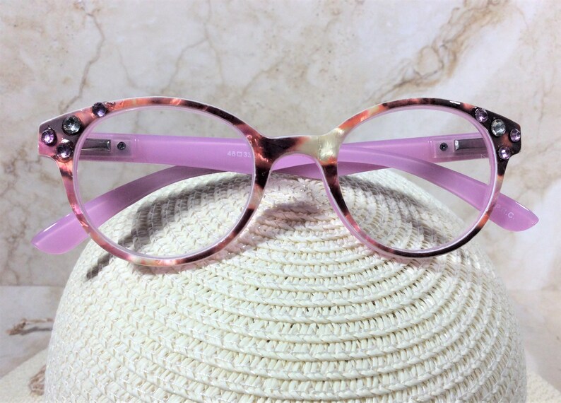 May include: Round-framed eyeglasses with a pink and tortoiseshell pattern. The frames have clear lenses and are accented with rhinestones on the upper corners. The temples are a solid pink color. The glasses are resting on a white, woven surface.
