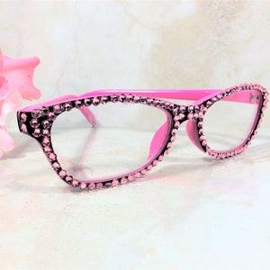 Pu&ograve; includere: Occhiali rosa con finiture di strass. Le montature sono rosa acceso, gli strass sono rosa pi&ugrave; chiaro. Gli occhiali hanno una classica forma a occhi di gatto. Lo sfondo &egrave; sfocato, con un fiore rosa.