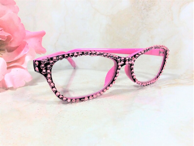 Pu&ograve; includere: Occhiali rosa con finiture di strass. Le montature sono rosa acceso, gli strass sono rosa pi&ugrave; chiaro. Gli occhiali hanno una classica forma a occhi di gatto. Lo sfondo &egrave; sfocato, con un fiore rosa.