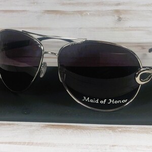 May include: Silver aviator sunglasses with dark lenses. The sunglasses have a decorative silver accent on the temples and the text "Maid of Honor" on the lens.