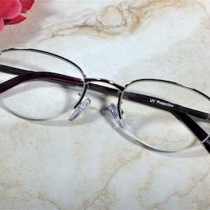May include: Silver-rimmed spectacles with a half-frame design. The arms are a deep purple, and the ends are decorated with iridescent rhinestones. The lenses have UV Protection. Made in China.