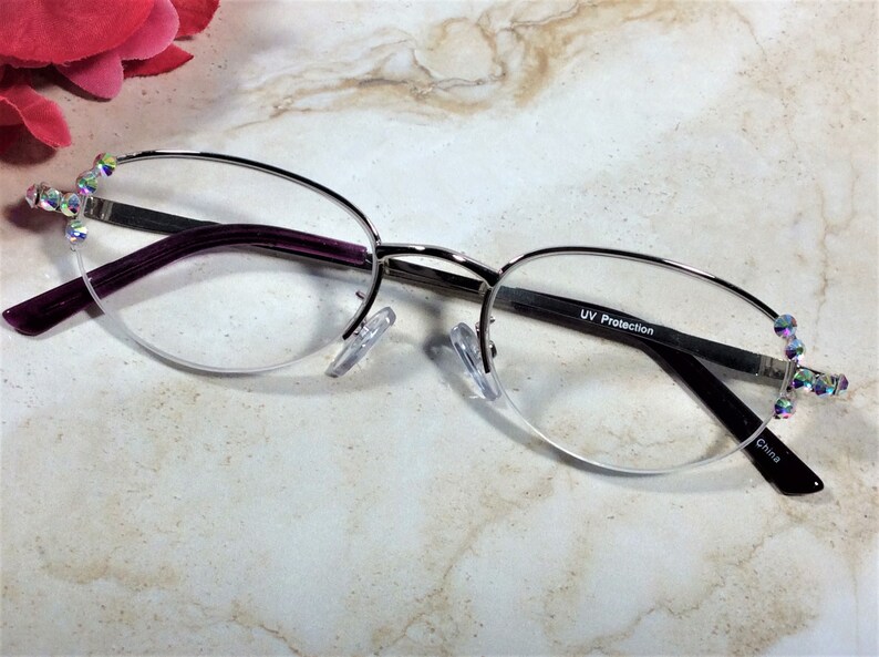 May include: Silver-rimmed spectacles with a half-frame design. The arms are a deep purple, and the ends are decorated with iridescent rhinestones. The lenses have UV Protection. Made in China.
