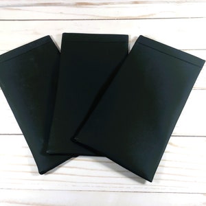 Puede incluir: Tres bolsas de tela negras, posiblemente para gafas u otros art&iacute;culos peque&ntilde;os, est&aacute;n apiladas sobre una superficie de madera blanca.
