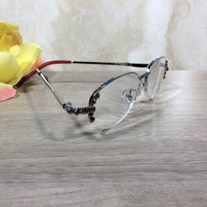 Puede incluir: Gafas con montura plateada, con una patilla roja y negra y un acento azul. Las gafas tienen un dise&ntilde;o sin montura con peque&ntilde;as decoraciones en la montura. Una flor amarilla est&aacute; en el fondo.