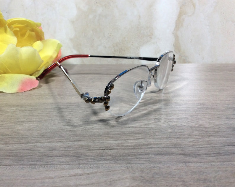 Puede incluir: Gafas con montura plateada, con una patilla roja y negra y un acento azul. Las gafas tienen un dise&ntilde;o sin montura con peque&ntilde;as decoraciones en la montura. Una flor amarilla est&aacute; en el fondo.