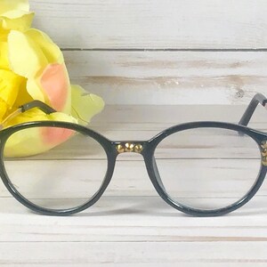 May include: Black eyeglasses featuring a rounded frame design with small gold-toned metallic studs embedded along the bridge and the outer corners of the rims. The temples are thin and gold-toned, connecting to the black front frame pieces.