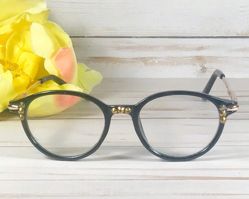 May include: Black eyeglasses featuring a rounded frame design with small gold-toned metallic studs embedded along the bridge and the outer corners of the rims. The temples are thin and gold-toned, connecting to the black front frame pieces.