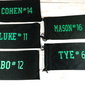 May include: Black drawstring pouches with green text. The pouches display names and numbers: COHEN #14, LUKE #11, BO #12, MASON #16, and TYE #64. Each pouch has a black drawstring closure.