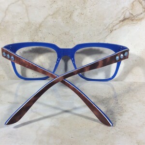 May include: Blue-framed eyeglasses with brown patterned temples and clear lenses. The temples are adorned with three silver-colored studs. The glasses are set against a neutral background.
