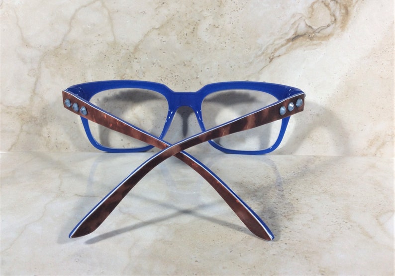 May include: Blue-framed eyeglasses with brown patterned temples and clear lenses. The temples are adorned with three silver-colored studs. The glasses are set against a neutral background.