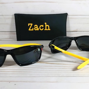 Op de afbeelding: Zwarte zonnebril met gele monturen en een zwarte etui met de naam "Zach" erop geborduurd.