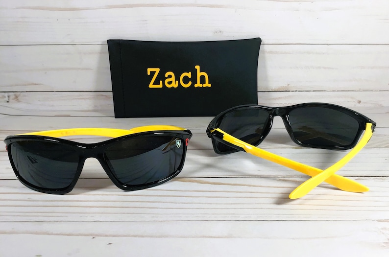 Op de afbeelding: Zwarte zonnebril met gele monturen en een zwarte etui met de naam "Zach" erop geborduurd.