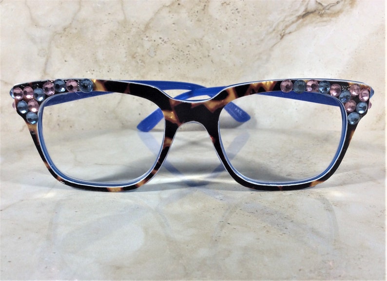May include: Eyeglasses with a tortoiseshell pattern frame and blue accents. The top of the frame is embellished with pink and blue rhinestones. The lenses are clear. The glasses are designed for vision correction.