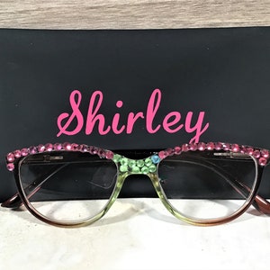 May include: Cat-eye style reading glasses with a gradient frame, adorned with pink rhinestones along the top rim. The glasses rest in front of a black case with the name "Shirley" in pink script.