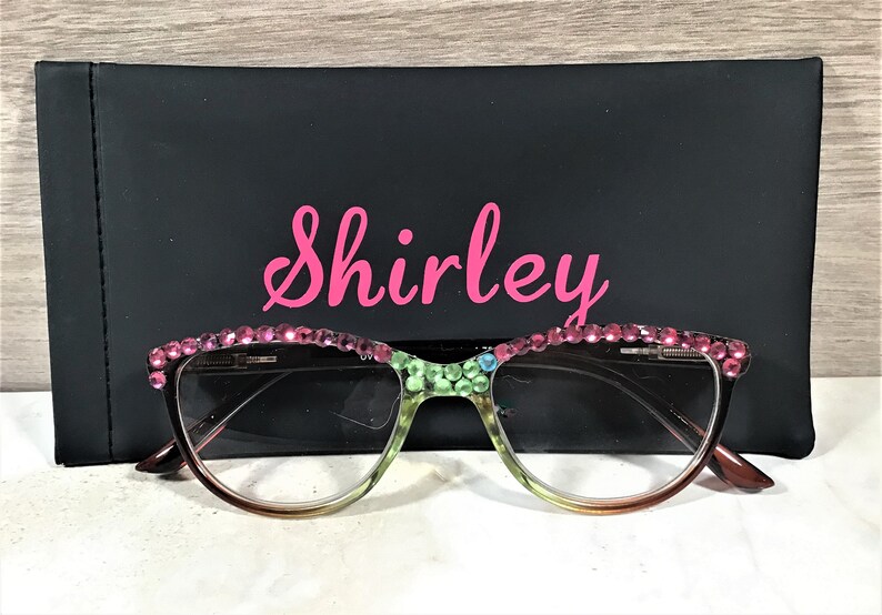 May include: Cat-eye style reading glasses with a gradient frame, adorned with pink rhinestones along the top rim. The glasses rest in front of a black case with the name "Shirley" in pink script.
