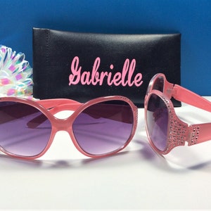 May include: Pink sunglasses with rhinestones on the arms and a black case with the name "Gabrielle" printed on it.