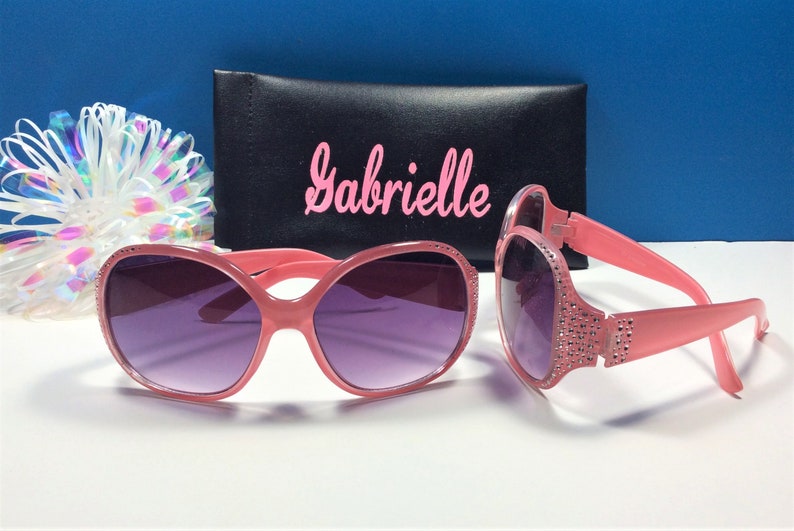 May include: Pink sunglasses with rhinestones on the arms and a black case with the name "Gabrielle" printed on it.