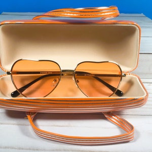May include: Heart-shaped orange sunglasses with gold frames inside an open matching case. The case and handle are orange with white stripes. The sunglasses have a UV Protection label.