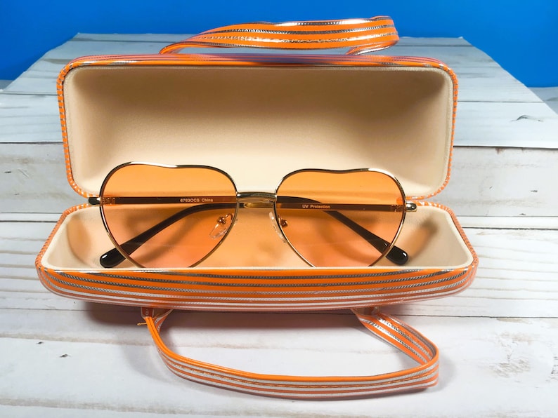 May include: Heart-shaped orange sunglasses with gold frames inside an open matching case. The case and handle are orange with white stripes. The sunglasses have a UV Protection label.