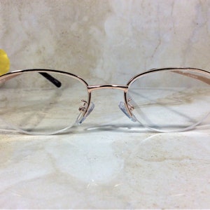 Pu&ograve; includere: Occhiali color oro rosa con lenti trasparenti. Le montature hanno una forma ovale delicata e sono impreziosite da piccole gemme viola sulle aste. Gli occhiali sono appoggiati su uno sfondo morbido e neutro.
