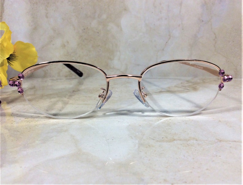 Pu&ograve; includere: Occhiali color oro rosa con lenti trasparenti. Le montature hanno una forma ovale delicata e sono impreziosite da piccole gemme viola sulle aste. Gli occhiali sono appoggiati su uno sfondo morbido e neutro.
