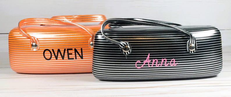May include: Two striped cosmetic bags. One is orange with the name "OWEN" in black. The other is black and white with the name "Anna" in pink. Both bags have a handle and silver hardware.
