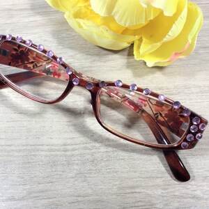 May include: Brown-framed reading glasses with a floral pattern and embellished with purple rhinestones. The glasses have a rectangular shape and are positioned near a yellow flower.