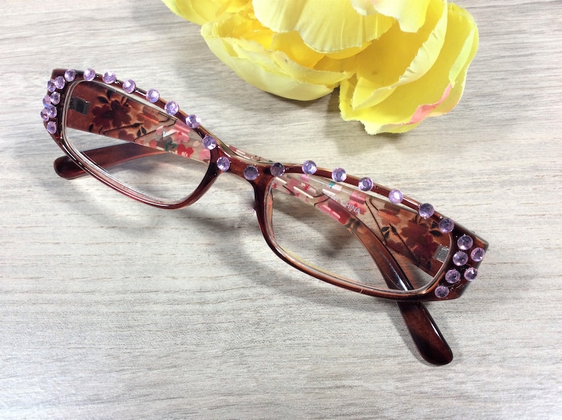 May include: Brown-framed reading glasses with a floral pattern and embellished with purple rhinestones. The glasses have a rectangular shape and are positioned near a yellow flower.