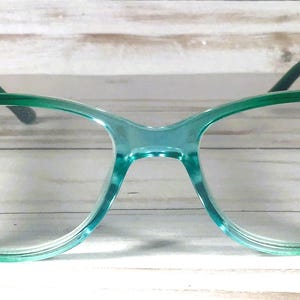 May include: Cat-eye glasses with a translucent teal frame that transitions to a darker green at the outer edges. The temples feature three small, round, light pink decorative rhinestones on each side near the hinges.