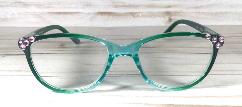 May include: Cat-eye glasses with a translucent teal frame that transitions to a darker green at the outer edges. The temples feature three small, round, light pink decorative rhinestones on each side near the hinges.