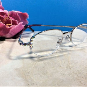 May include: Silver-framed spectacles with clear lenses. The frames have a decorative design with iridescent rhinestones at the temples. The glasses have a purple and silver arm. A pink flower is in the background.