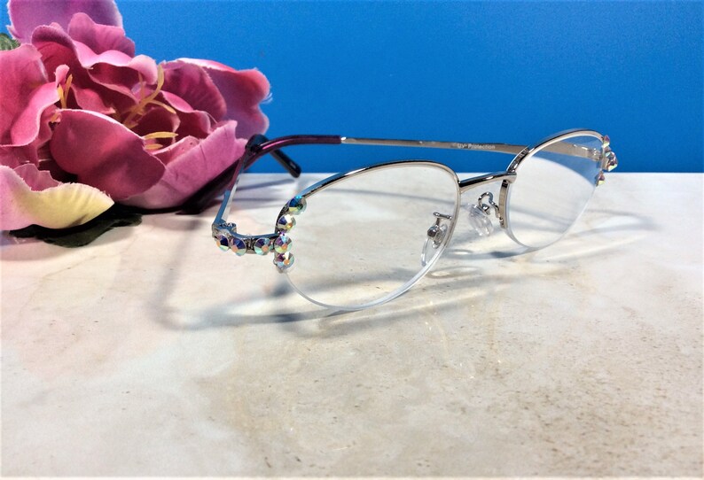 May include: Silver-framed spectacles with clear lenses. The frames have a decorative design with iridescent rhinestones at the temples. The glasses have a purple and silver arm. A pink flower is in the background.