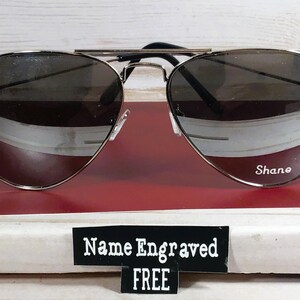 May include: Aviator-style sunglasses with a silver frame and dark lenses. The name "Shane" is engraved on the right lens. A sign below the sunglasses reads "Name Engraved FREE".