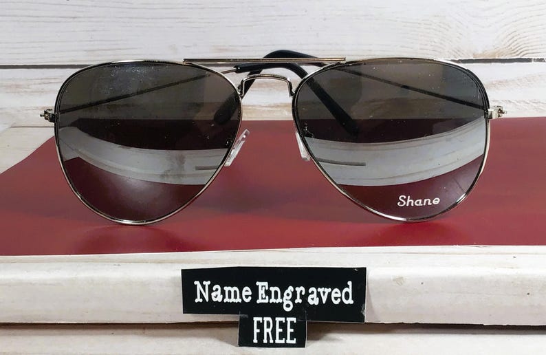May include: Aviator-style sunglasses with a silver frame and dark lenses. The name "Shane" is engraved on the right lens. A sign below the sunglasses reads "Name Engraved FREE".
