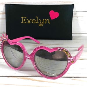 May include: Pink heart-shaped sunglasses with rhinestones and a crown detail. The sunglasses come with a black case that says "Evelyn" in gold lettering.