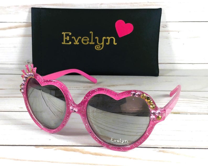 May include: Pink heart-shaped sunglasses with rhinestones and a crown detail. The sunglasses come with a black case that says "Evelyn" in gold lettering.