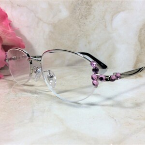 May include: Silver-rimmed spectacles with a semi-rimless design. The frames feature a decorative cluster of purple gemstones on the outer corners. The lenses are clear, and the temples are silver with black accents.
