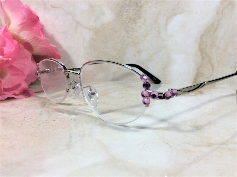 May include: Silver-rimmed spectacles with a semi-rimless design. The frames feature a decorative cluster of purple gemstones on the outer corners. The lenses are clear, and the temples are silver with black accents.