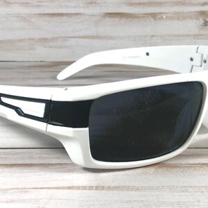Pu&ograve; includere: Occhiali da sole bianchi con lenti scure. Gli occhiali da sole presentano un design sportivo con una montatura bianca spessa e un accento nero sulle aste. Le lenti sono scure, offrendo ombra e protezione solare.