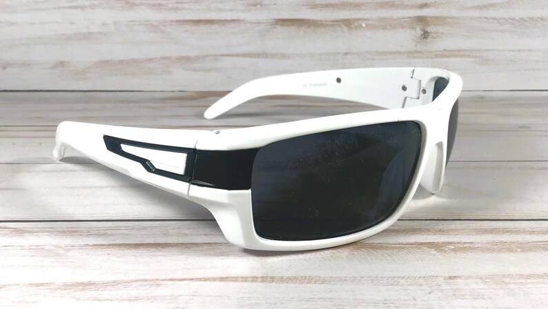 Pu&ograve; includere: Occhiali da sole bianchi con lenti scure. Gli occhiali da sole presentano un design sportivo con una montatura bianca spessa e un accento nero sulle aste. Le lenti sono scure, offrendo ombra e protezione solare.