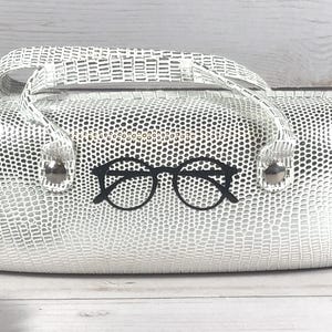 May include: A white glasses case with a textured, reptile-like pattern. The case has a handle and a black glasses graphic on the front. The case is on a wooden surface.