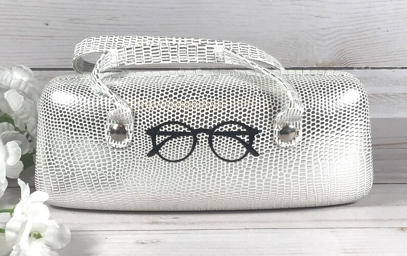 May include: A white glasses case with a textured, reptile-like pattern. The case has a handle and a black glasses graphic on the front. The case is on a wooden surface.
