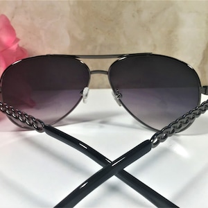 Peut inclure: Lunettes de soleil aviateur noires avec un d&eacute;tail de cha&icirc;ne sur les branches. Les verres sont d&eacute;grad&eacute;s, passant du fonc&eacute; au clair.