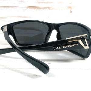 May include: Black sunglasses with dark lenses. The sunglasses have a sporty design with a thick frame and arms. The arms feature a silver accent and the text "XLOOP". The sunglasses are angled, showing the details of the frame.