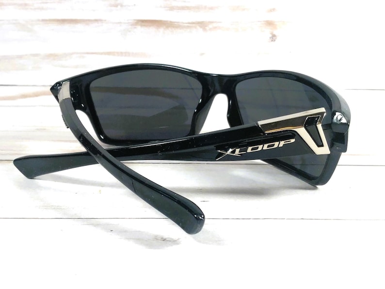 May include: Black sunglasses with dark lenses. The sunglasses have a sporty design with a thick frame and arms. The arms feature a silver accent and the text "XLOOP". The sunglasses are angled, showing the details of the frame.