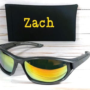 May include: Gray sunglasses with mirrored lenses reflecting yellow and orange hues. A black case with the name "Zach" in yellow lettering is behind the sunglasses. The sunglasses have a sporty design.