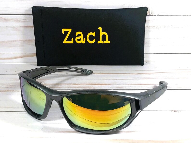 May include: Gray sunglasses with mirrored lenses reflecting yellow and orange hues. A black case with the name "Zach" in yellow lettering is behind the sunglasses. The sunglasses have a sporty design.