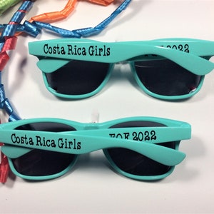 Op de afbeelding: Twee paar turquoise blauwe zonnebrillen met zwarte glazen. Het bovenste paar heeft de tekst "Costa Rica Girls" op het montuur. Het onderste paar heeft de tekst "TOF 2022" op het montuur.